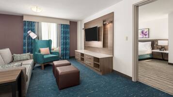 A hotel room with a sofa, ottoman, TV, desk, and a bed with a nightstand.