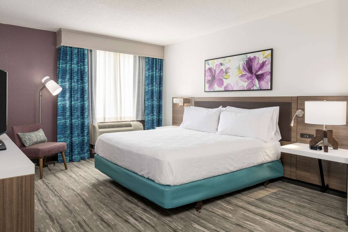 Habitación de hotel con cama, escritorio, silla, lámpara y ventana con cortinas.