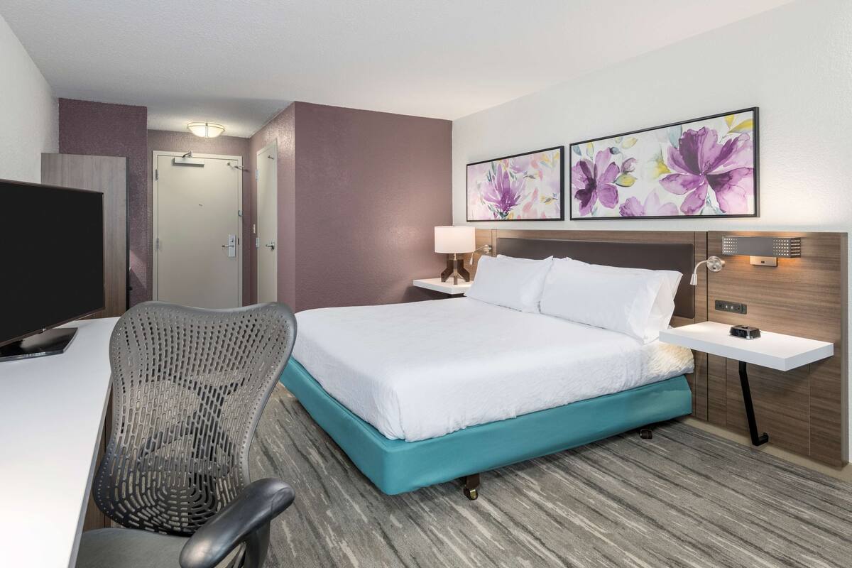 Una habitación de hotel con una cama, un escritorio con silla, televisión y obras de arte en la pared.