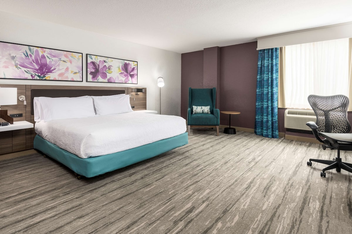 Habitación de hotel con una cama grande, un escritorio con lámpara, una silla y dos cuadros colgados en la pared.