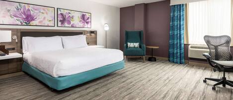A hotel room with a large bed, a desk with a lamp, a chair, and two wall-mounted paintings.