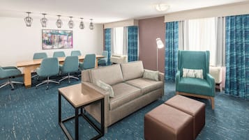 A hotel room with a sofa, ottoman, a small table, a desk, and chairs. There is a large window with curtains, a floor lamp, and a ceiling light fixture.