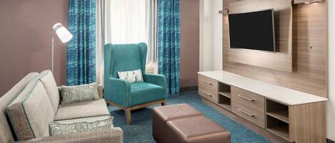 Habitación de hotel moderna con sofá, sillón, reposapiés, televisión y escritorio con cajones.