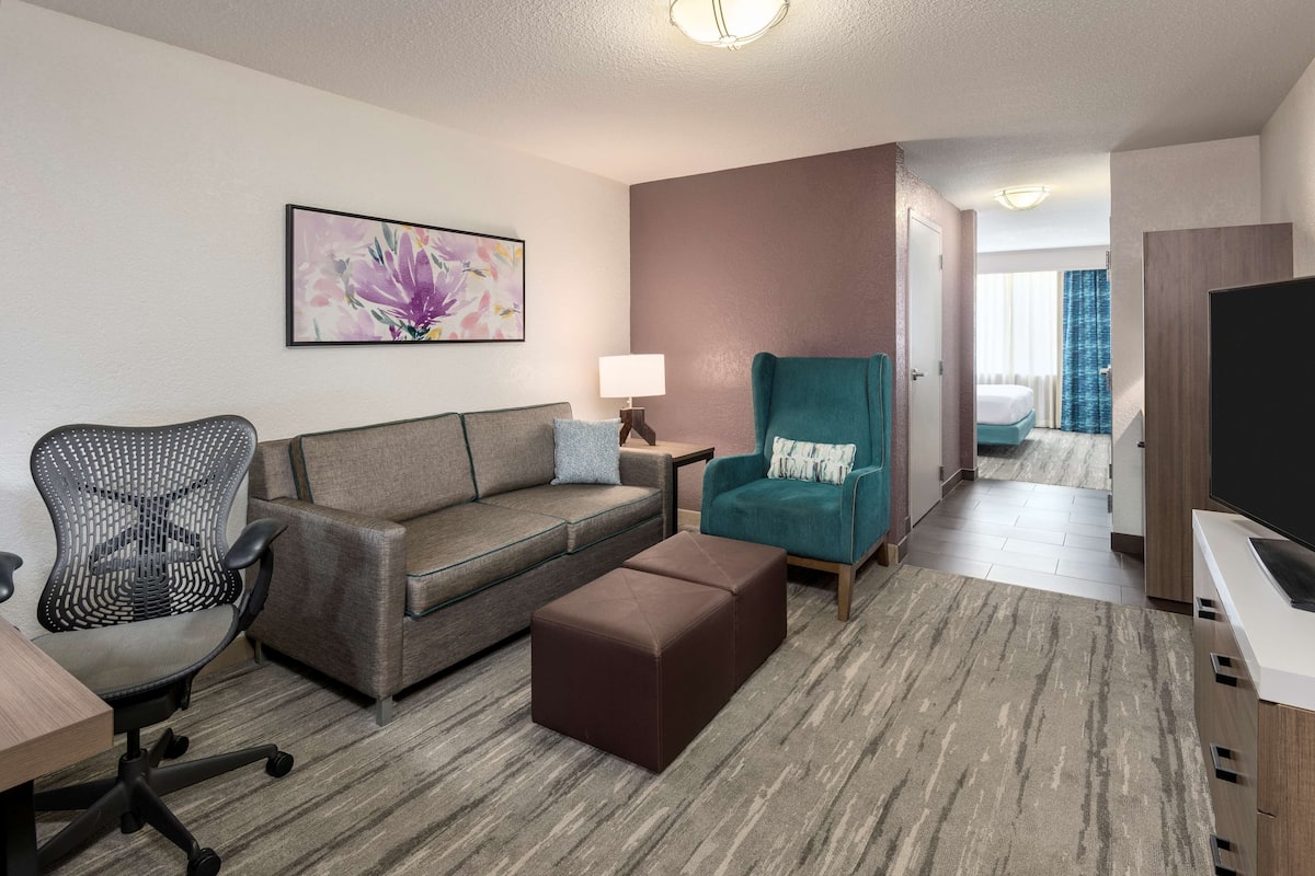 Una habitación de hotel moderna con sofá, sillón, reposapiés, escritorio, silla y televisor.