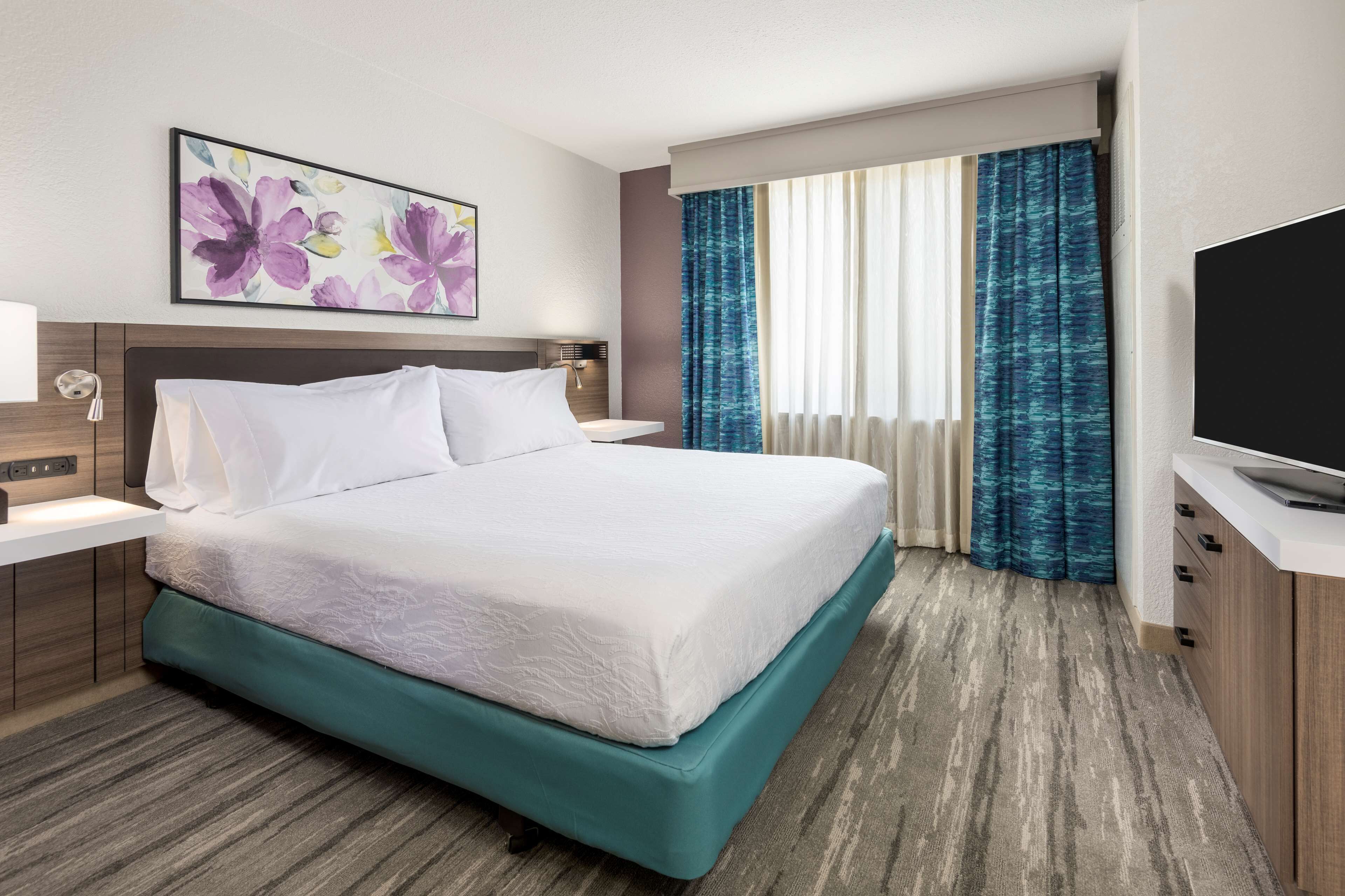 A hotel room with a large bed, a television, a painting, and a window with curtains.