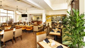 A spacious hotel dining area with multiple tables and chairs, a large plant, and a television on the wall. - Hilton Garden Inn Anderson (Anderson)