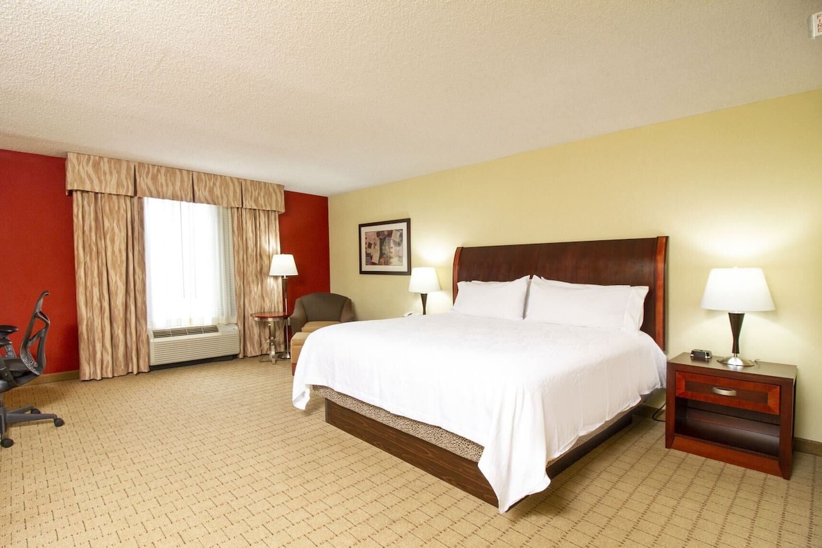 A hotel room with a bed, two bedside lamps, a chair, a desk, and a window with curtains.