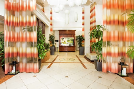 A hotel lobby with a reception desk, potted plants, and a tiled floor.
