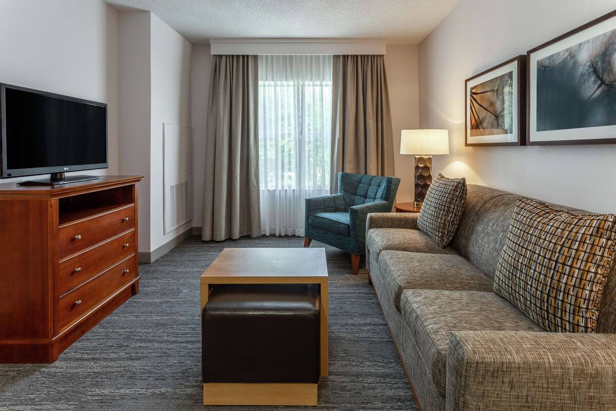 A hotel room with a flat-screen TV, a wooden dresser, a sofa, a small table, a chair, and a window with curtains.