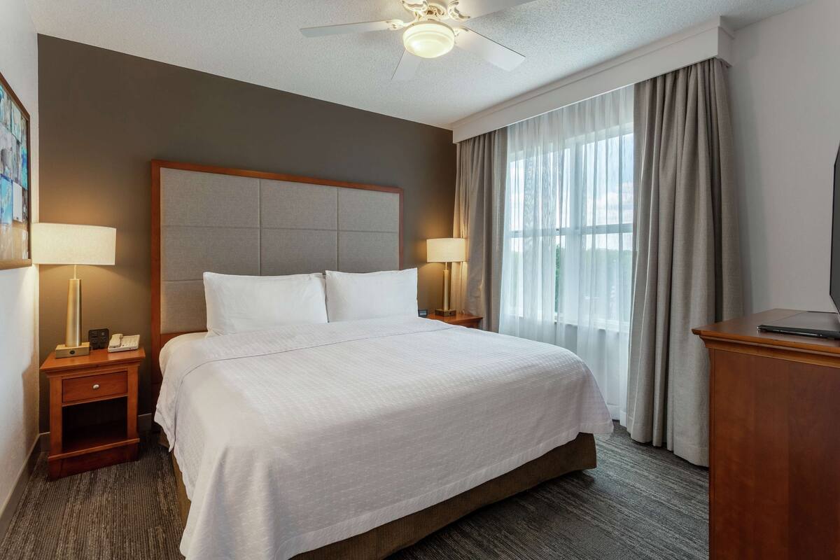 A hotel room with a large bed, two bedside tables with lamps, a ceiling fan, and a large window with curtains.
