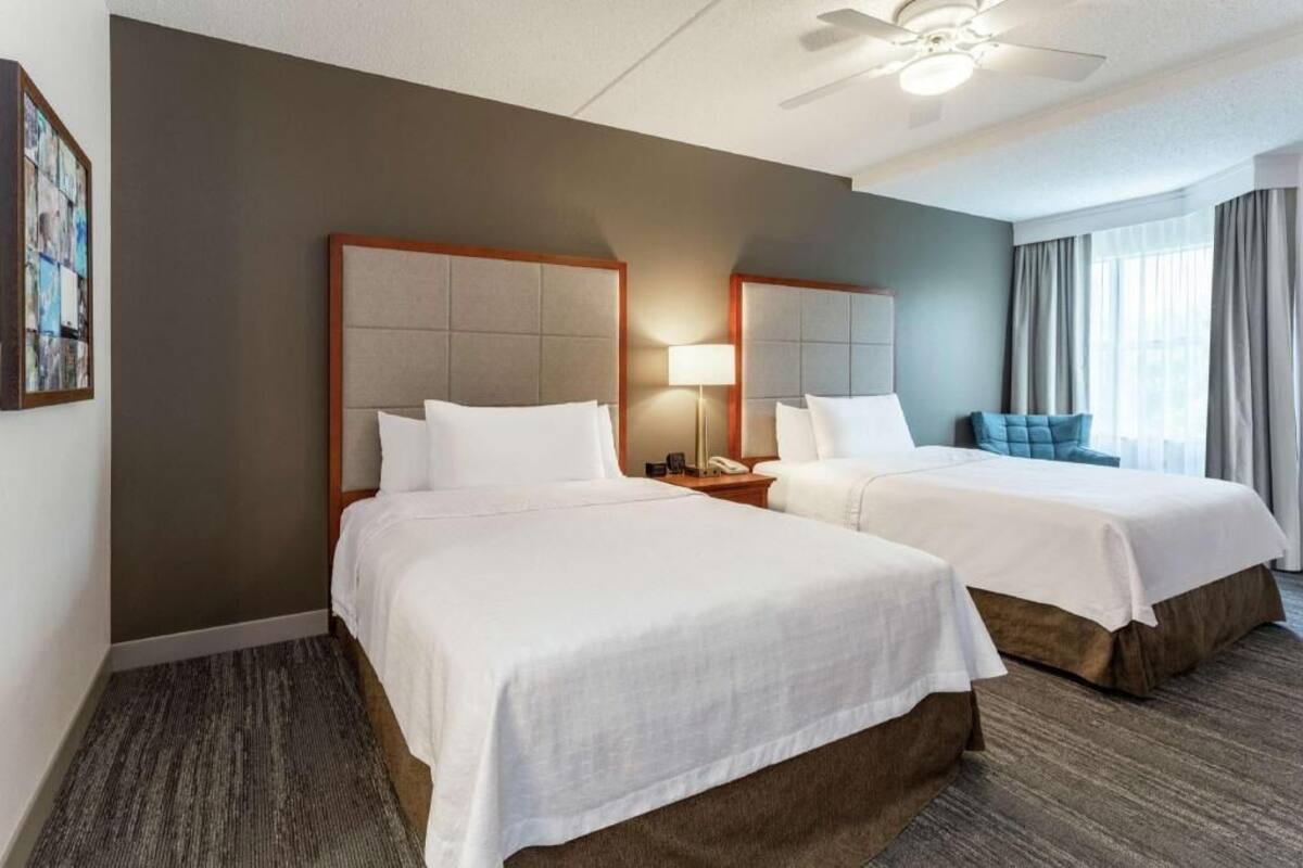 A hotel room with two beds, a ceiling fan, a bedside lamp, a telephone, and a window with curtains.