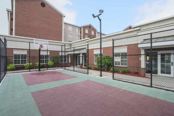 A basketball court with a fence, a basketball hoop, and a building with a brick facade in the background.