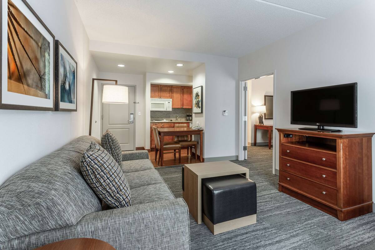 A modern living room with a gray sofa, a wooden coffee table, a TV on a wooden stand, and a kitchen in the background.