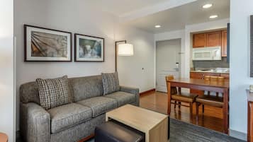 A modern living room with a gray sofa, a wooden coffee table, and a dining area with a wooden table and chairs. There are framed artworks on the wall and a lamp beside the sofa.