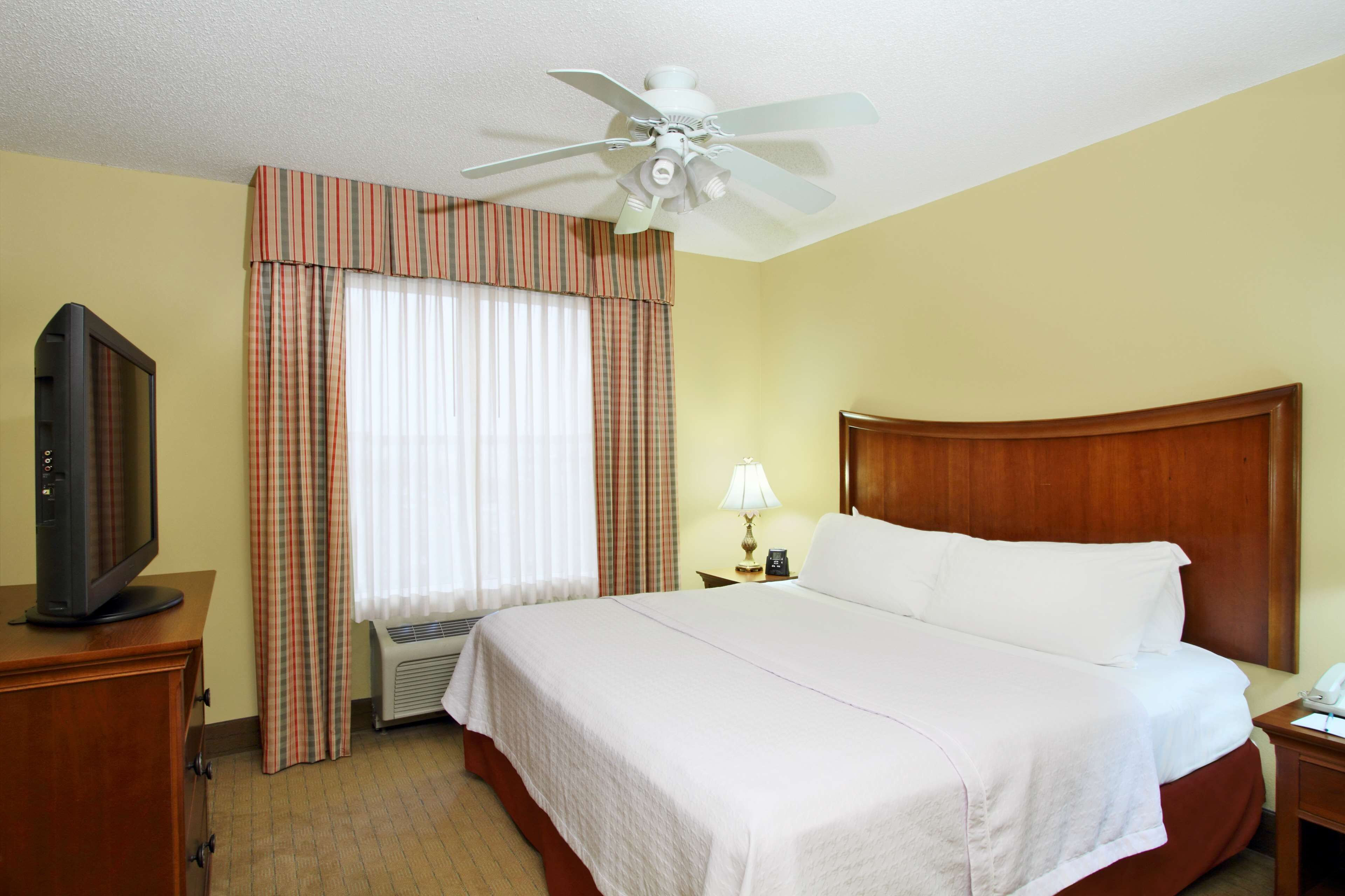 A hotel room with a bed, a television on a wooden dresser, a ceiling fan, and a window with curtains.