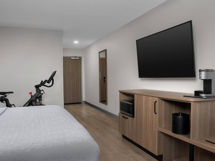 A hotel room with a bed, a wooden cabinet with a microwave, a bicycle, and a flat-screen TV on the wall.