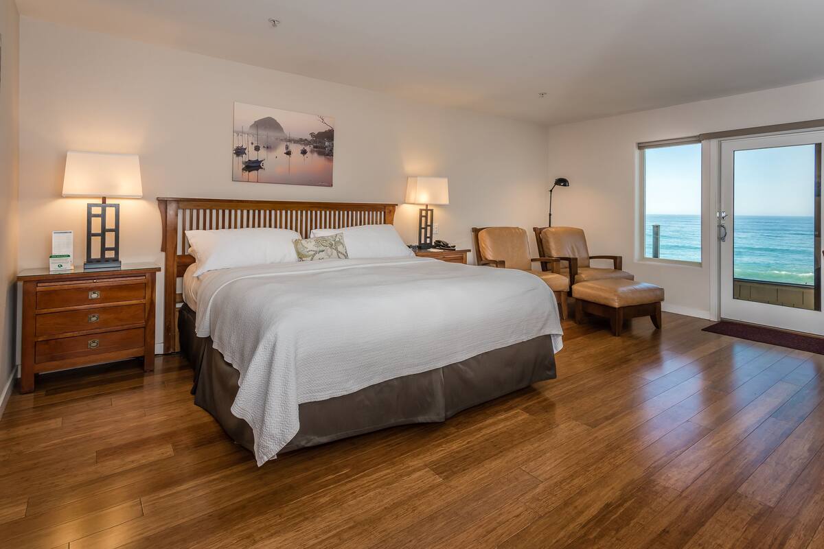 A hotel room with a bed, two chairs, a nightstand, a lamp, and a view of the sea.