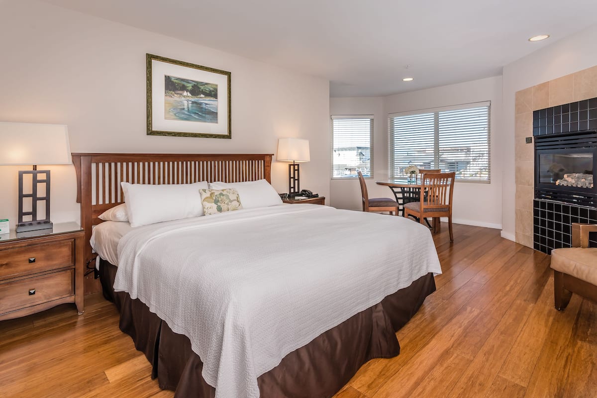 A bedroom with a large bed, wooden headboard, two bedside tables with lamps, a chair, a dining table with chairs, and a fireplace.