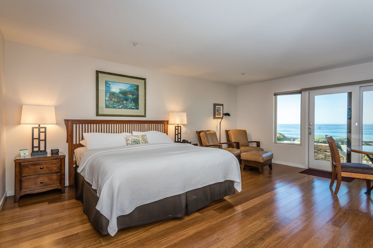 A bedroom with a large bed, two bedside tables, a chair, and a view of the ocean through a sliding glass door.