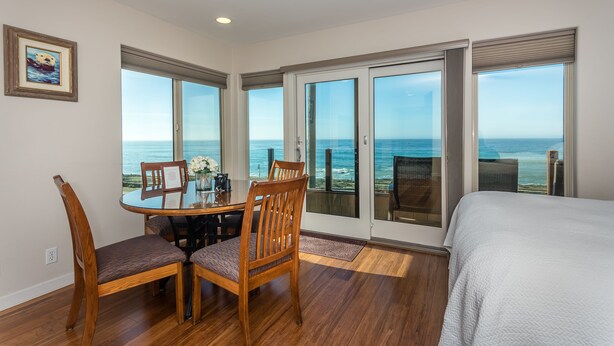 A bedroom with a large bed, a dining area with a table and chairs, and a view of the ocean through large sliding glass doors. - Little Sur Inn (Cambria)