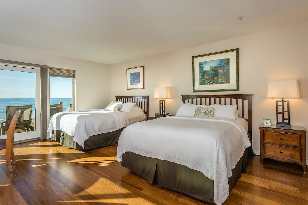 A hotel room with two beds, a wooden floor, a large window with a view of the sea, and a nightstand with a lamp.