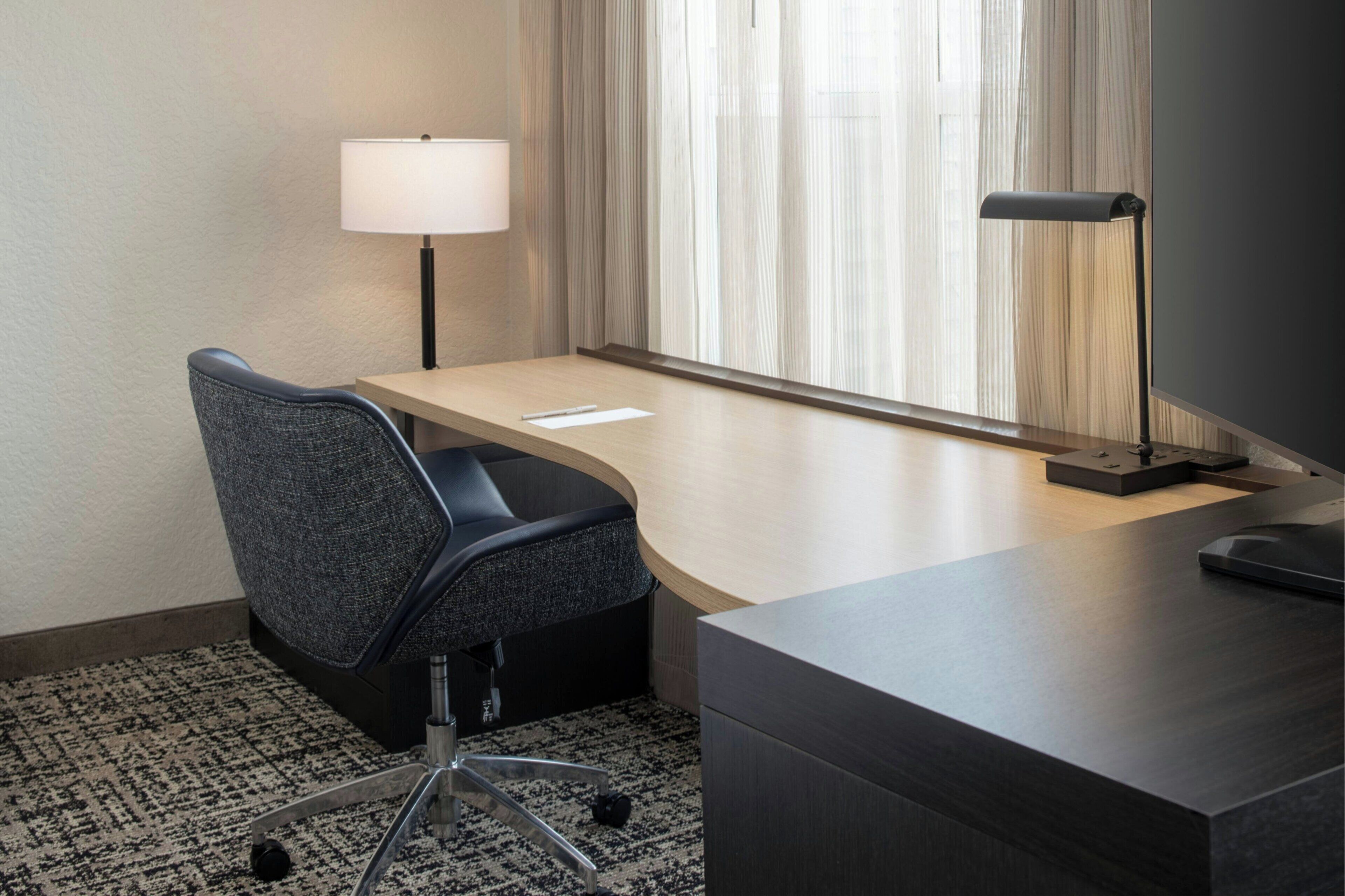 A hotel room with a desk, a chair, a lamp, and a window with curtains.