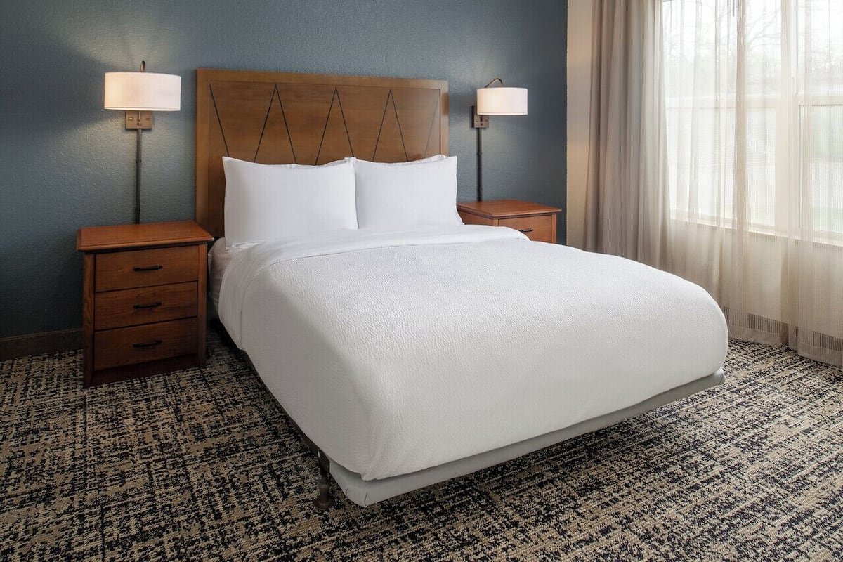Cama bem arrumada com cabeceira de madeira, duas mesas de cabeceira com abajures, tapete estampado e uma janela com cortinas leves.
