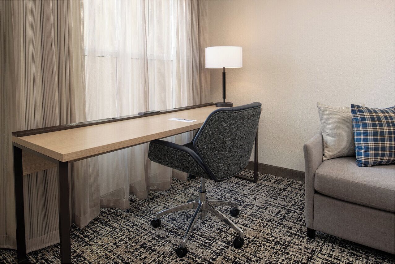 A hotel room with a desk, chair, lamp, and sofa.