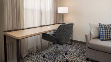 A hotel room with a desk, chair, lamp, and sofa.