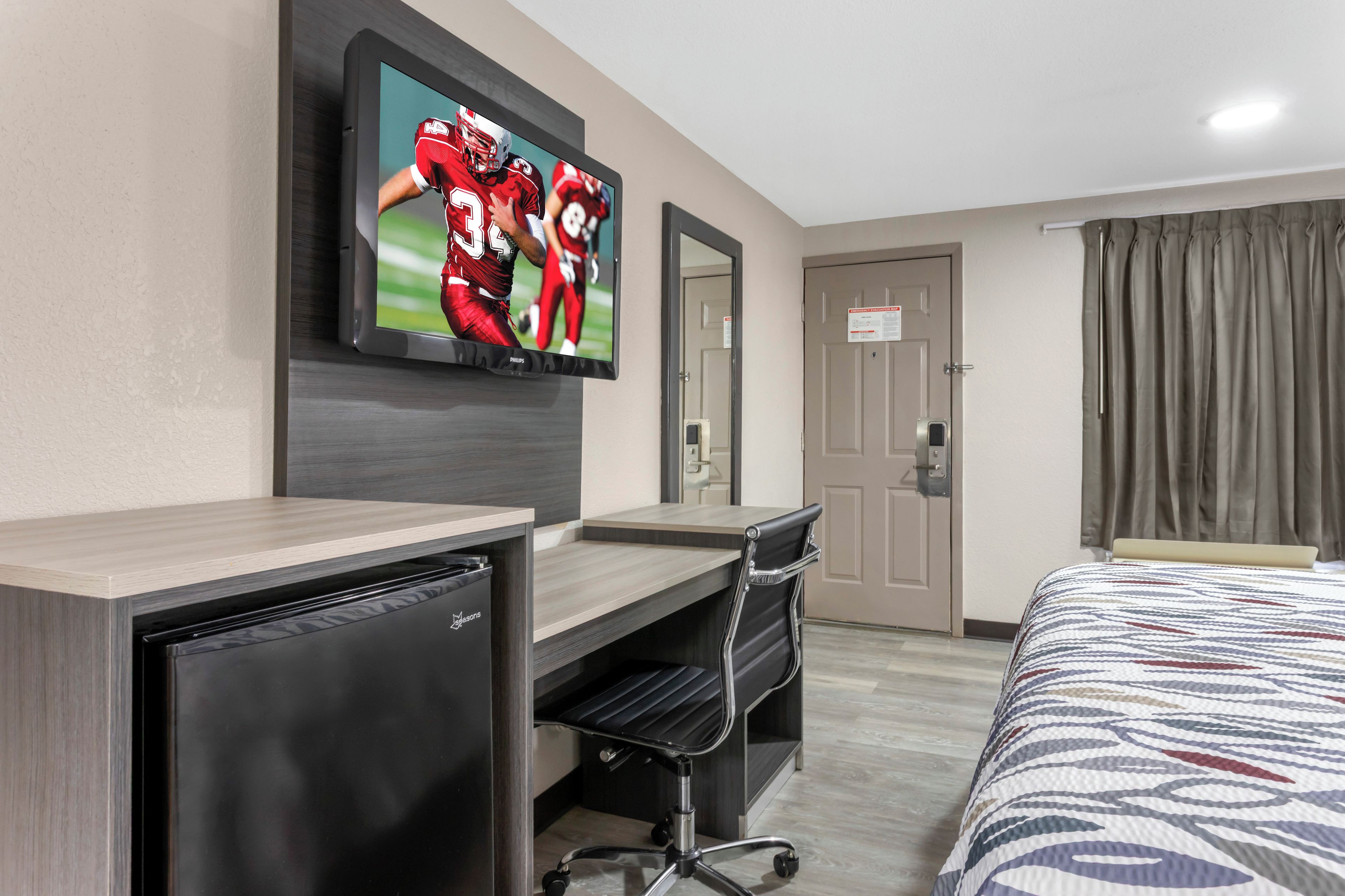 A hotel room with a bed, a desk with a computer, a television mounted on the wall, and a door with a sign.