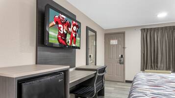 A hotel room with a bed, a desk with a computer, a television mounted on the wall, and a door with a sign.