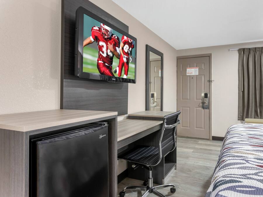 A hotel room with a bed, a desk with a computer, a television mounted on the wall, and a door with a sign.