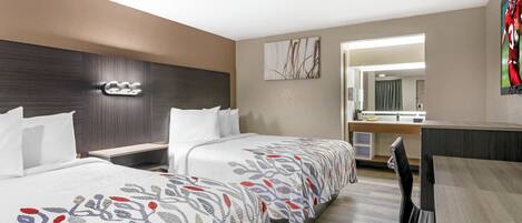 A hotel room with two beds, a desk, a mirror, and a bathroom visible through an open door.