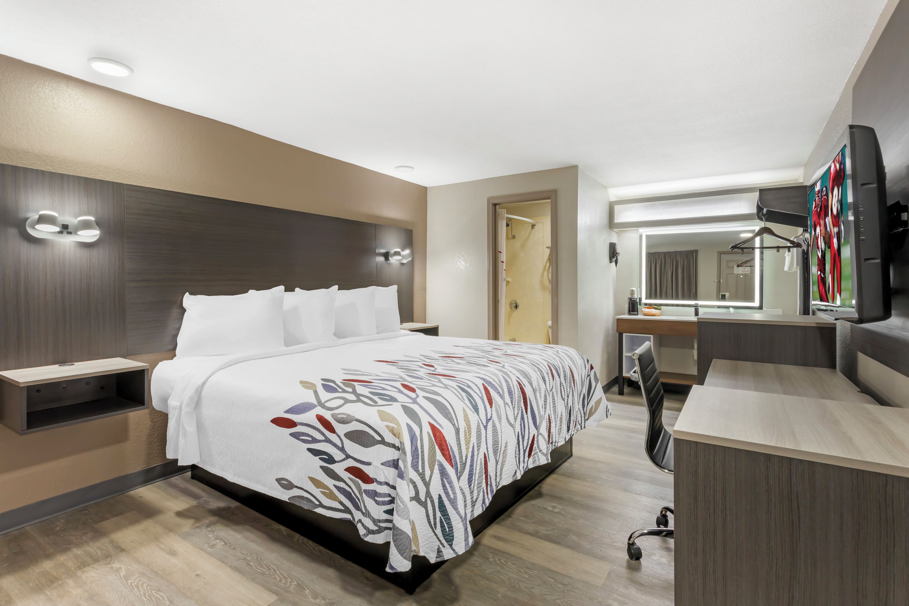 Habitación de hotel moderna con cama, mesitas de noche, escritorio, silla, espejo y televisor.