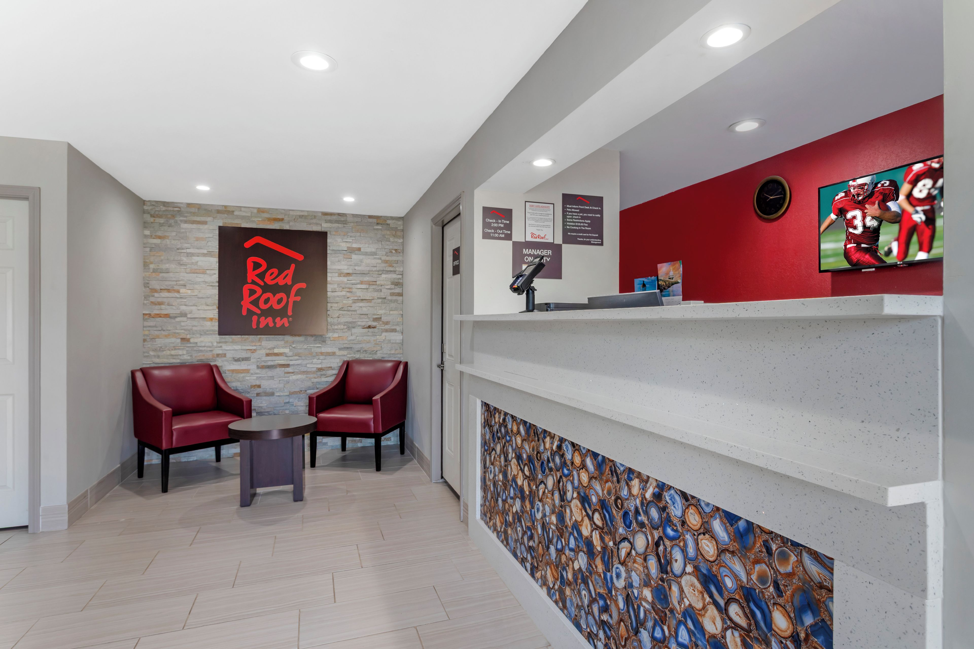 A reception area with a Red Roof Inn sign, two leather chairs, a reception desk, a wall clock, and a TV screen.