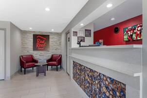 Un área de recepción con un letrero de Red Roof Inn, dos sillones de cuero, una mesa de recepción, un reloj de pared y una pantalla de televisión.
