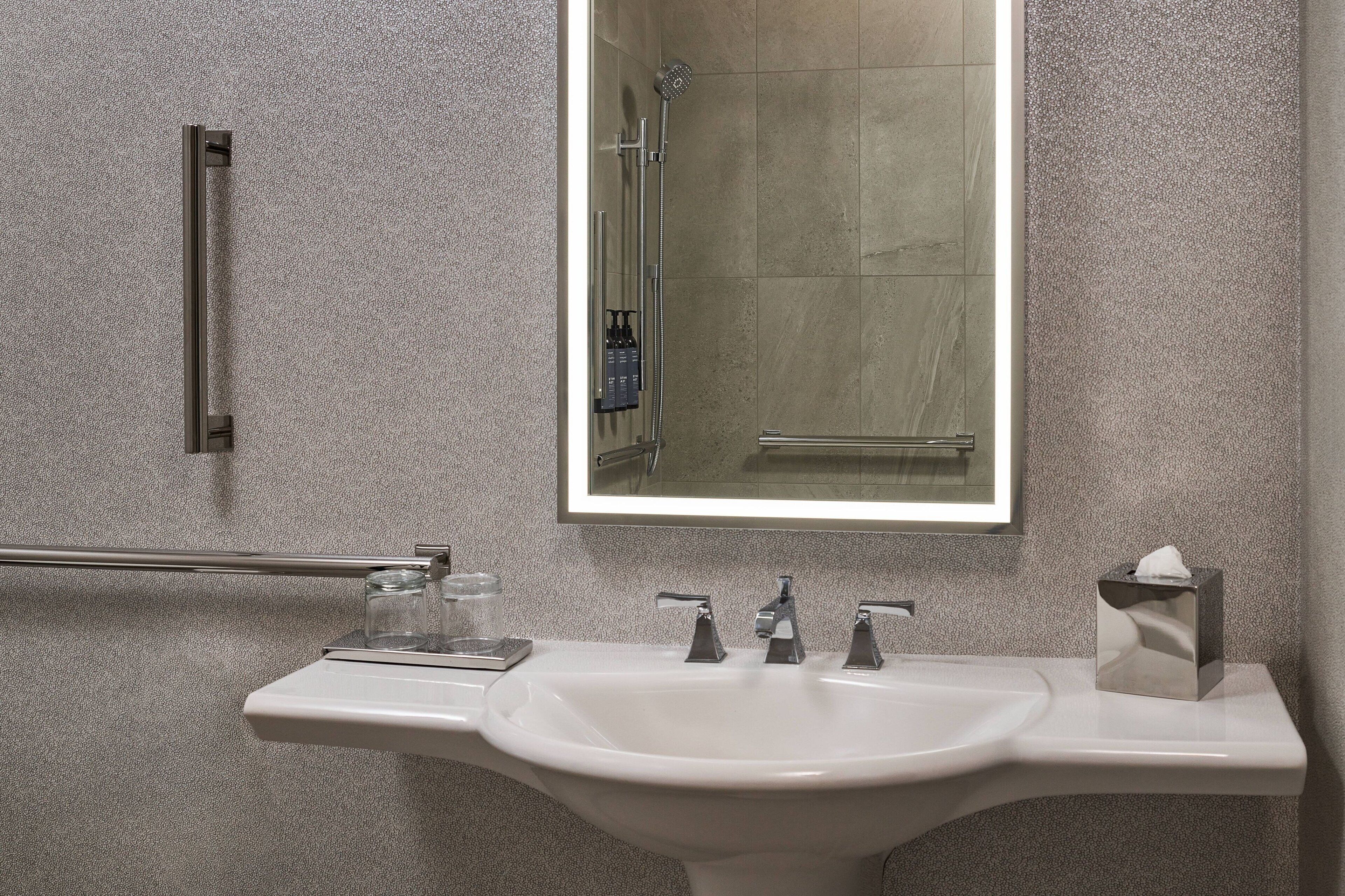 A bathroom with a white sink, two faucets, a mirror, a towel rack, and a glass of water on the counter.