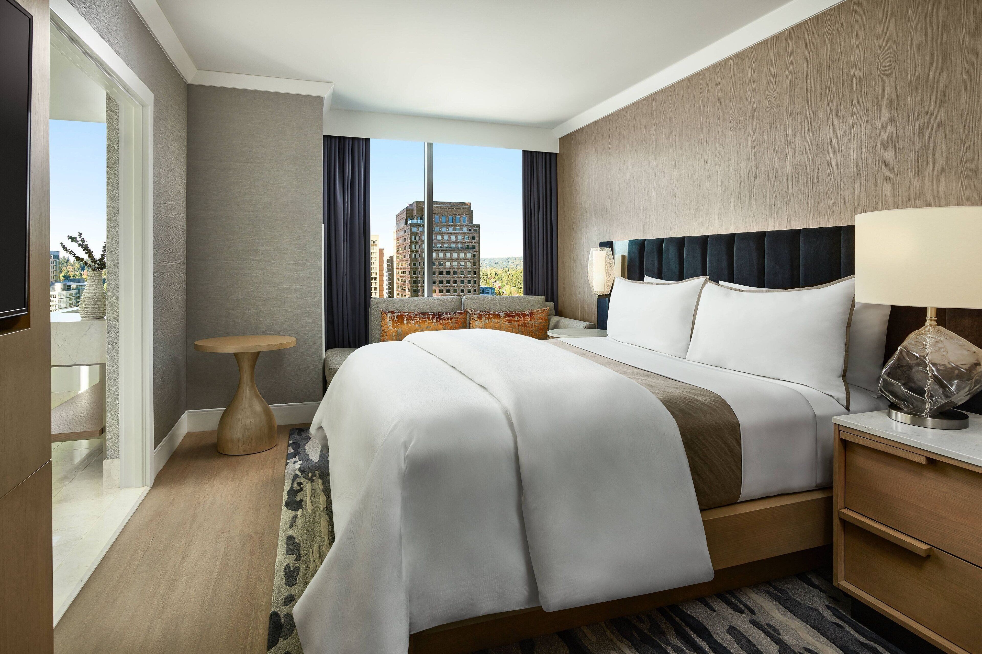 A hotel room with a large bed, a bedside table, a lamp, a small round table, and a view of buildings through the window.