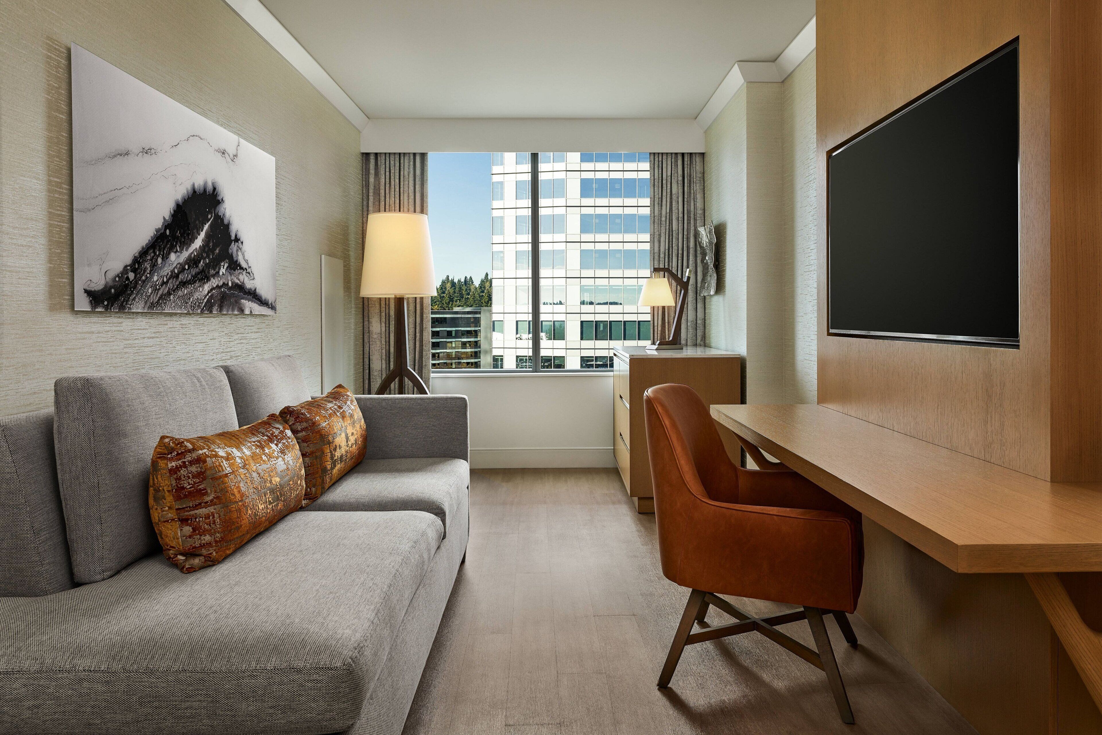 A modern hotel room with a grey sofa, a wooden desk, a large window with a view of buildings, and a black TV mounted on the wall.