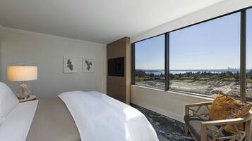 A hotel room with a bed, a chair, a TV, and a large window offering a city view.