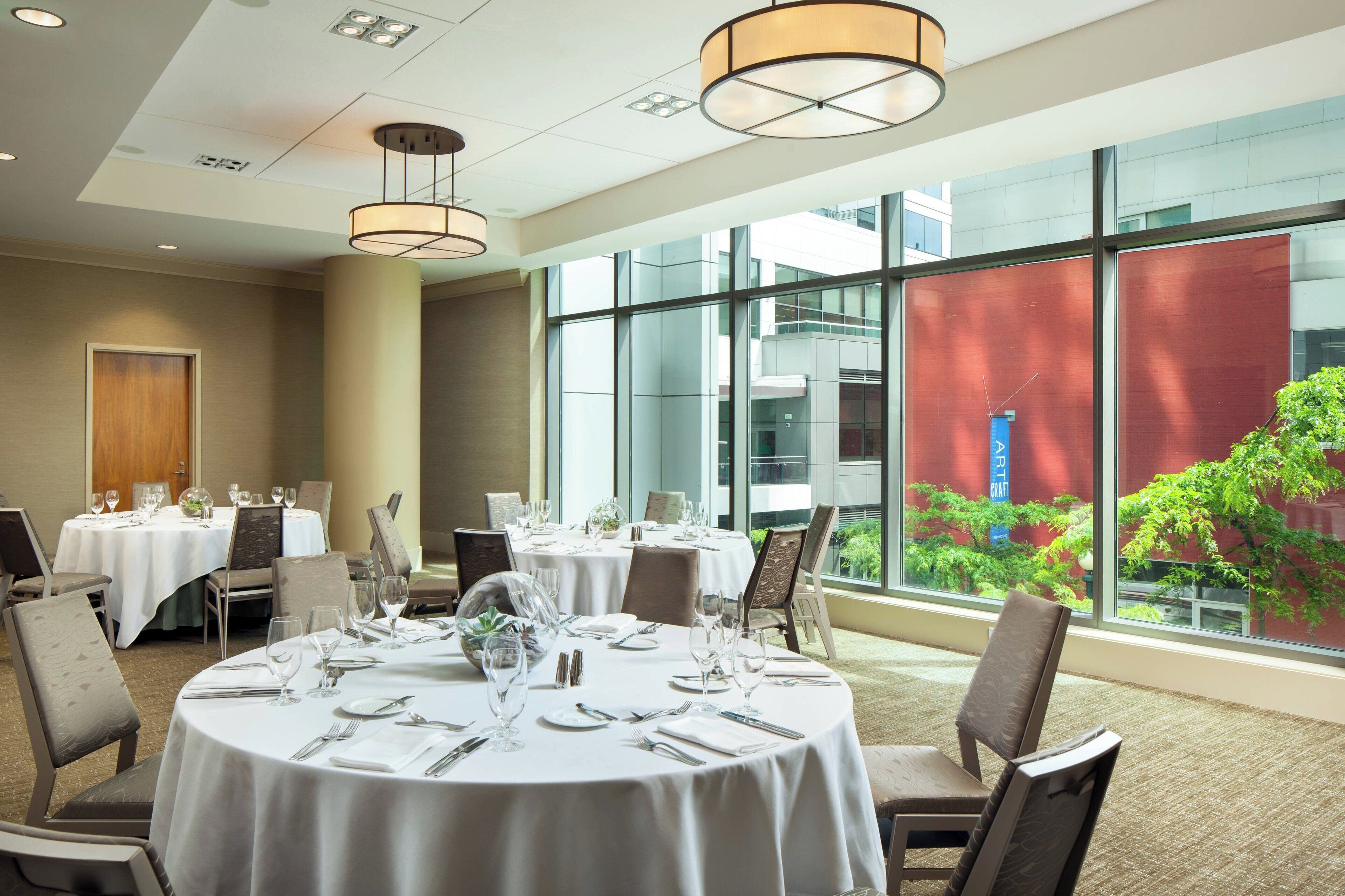 A dining area with round tables set for a meal, large windows with a view of greenery, and modern lighting fixtures.