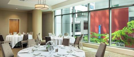 A dining area with round tables set for a meal, large windows with a view of greenery, and modern lighting fixtures.