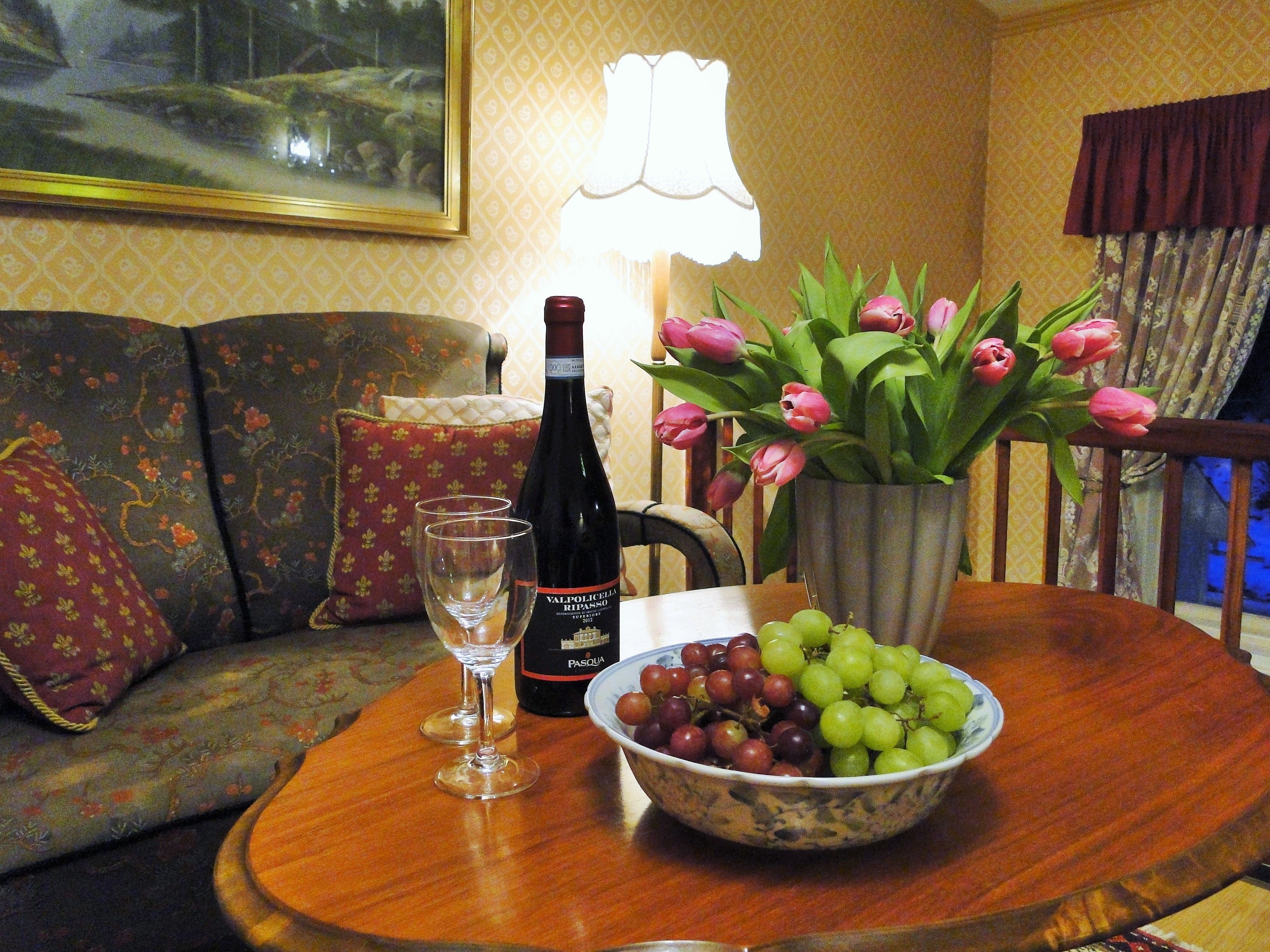 Una sala de estar con un sofá, una mesa con una botella de vino, un bol de uvas y un jarrón de tulipanes.