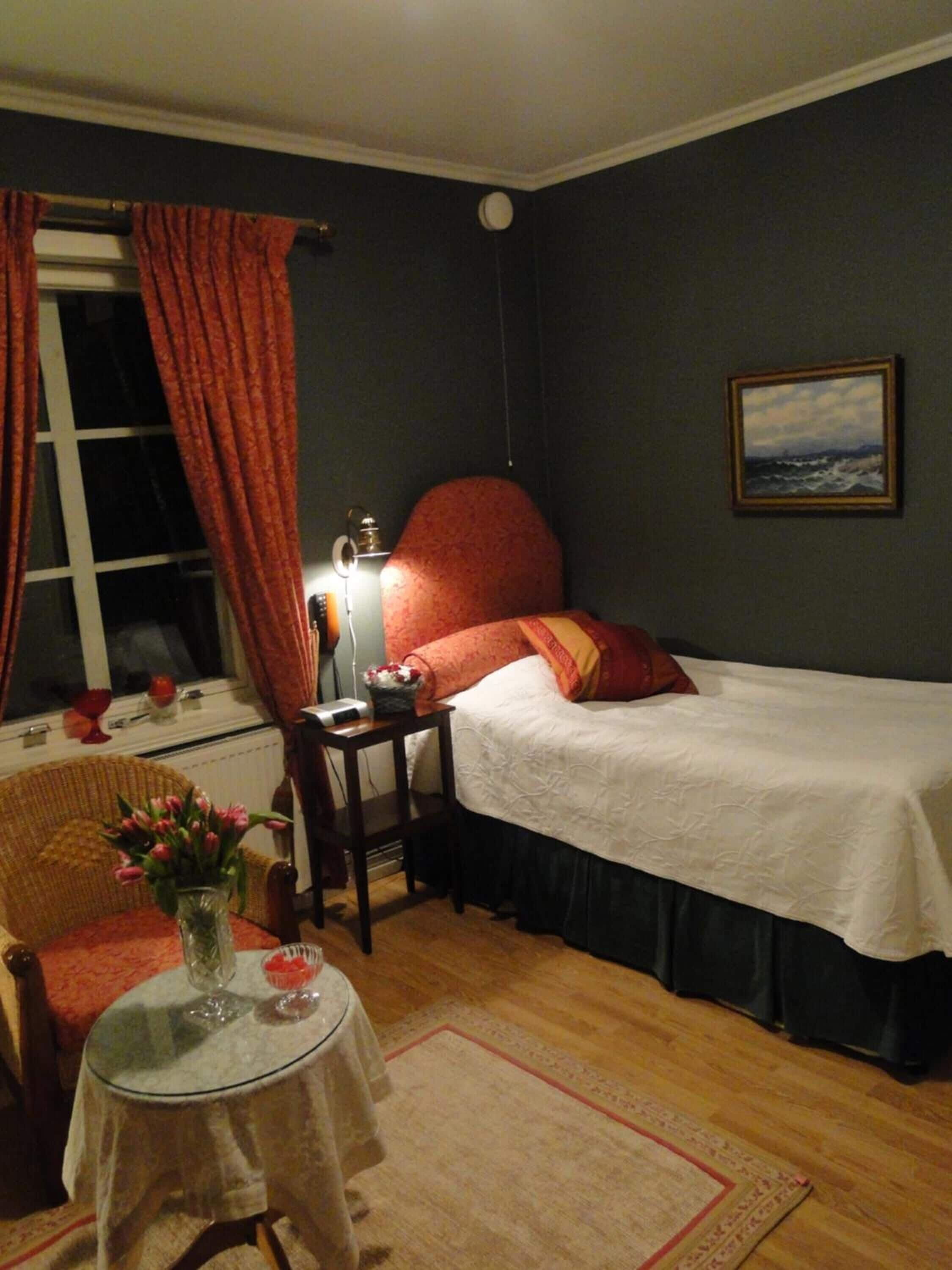 Une chambre avec un lit simple, une table de chevet avec une lampe, une chaise, une petite table avec un vase de fleurs et une fenêtre avec des rideaux.