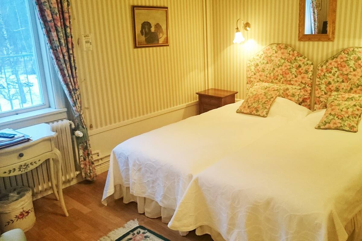 Una cama doble con cabecera y piecera adornados con motivos florales, una mesita de noche, un radiador, una ventana con cortinas y un cuadro en la pared.
