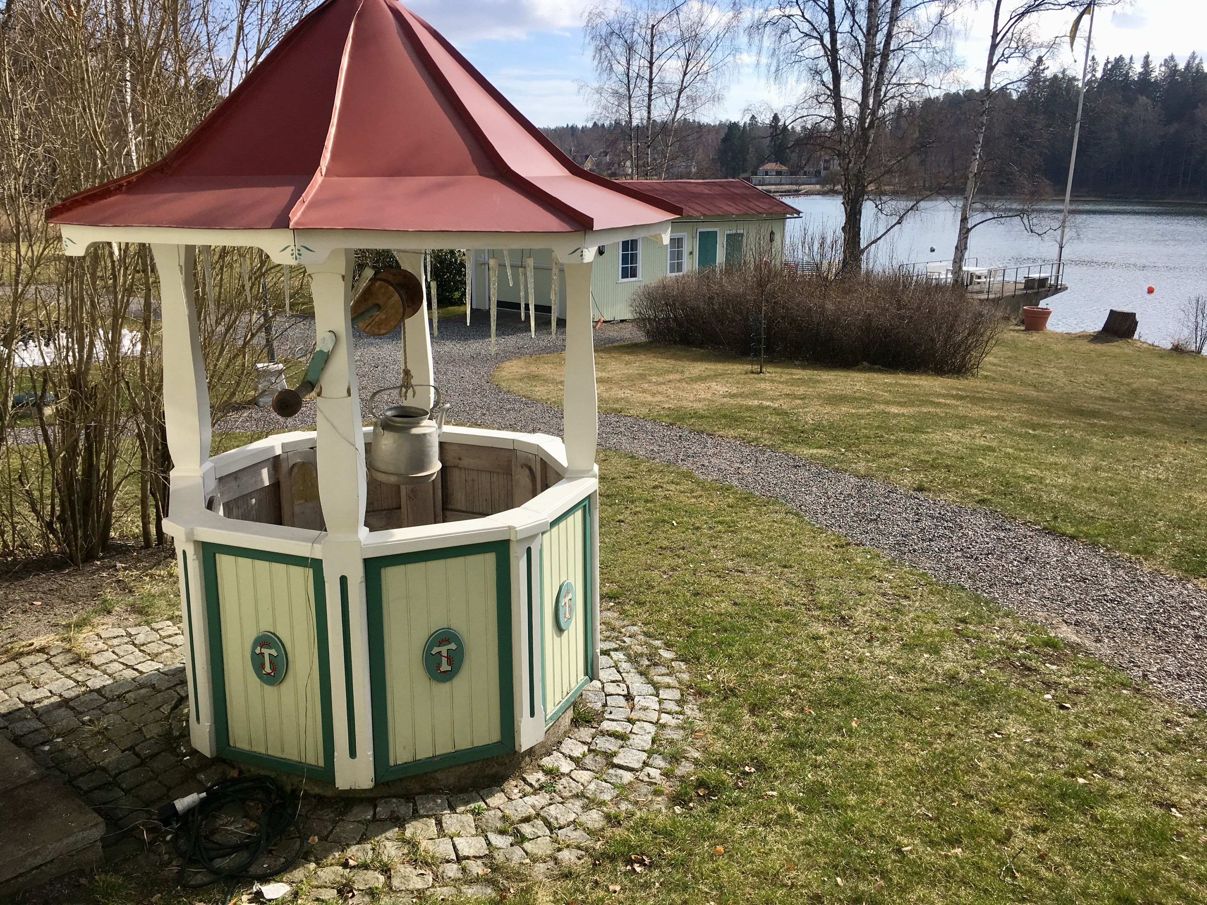 En lusthus med rött tak och gröna detaljer, omgivet av en stenlagd gång och en gräsyta, nära en sjö och en byggnad.