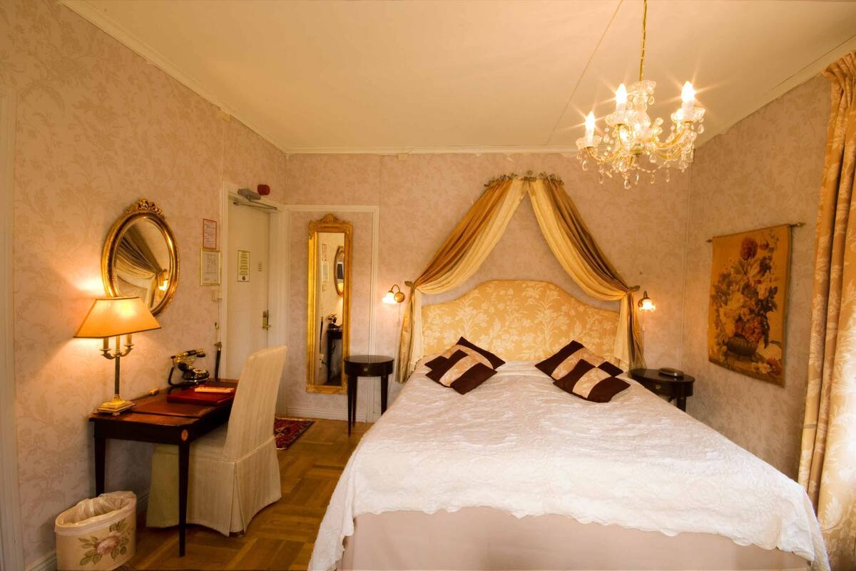 Un dormitorio con cama, escritorio, silla, espejo, candelabro y un cuadro en la pared.
