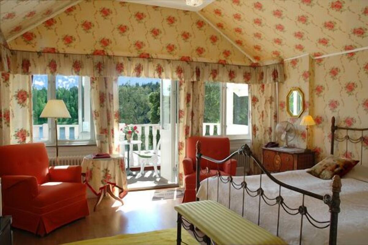 Una habitación con cama, un sillón rojo, una mesita y una ventana con vistas a zonas verdes.