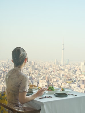 Una persona cenando en una mesa con vista a un paisaje urbano, que incluye una torre al fondo.