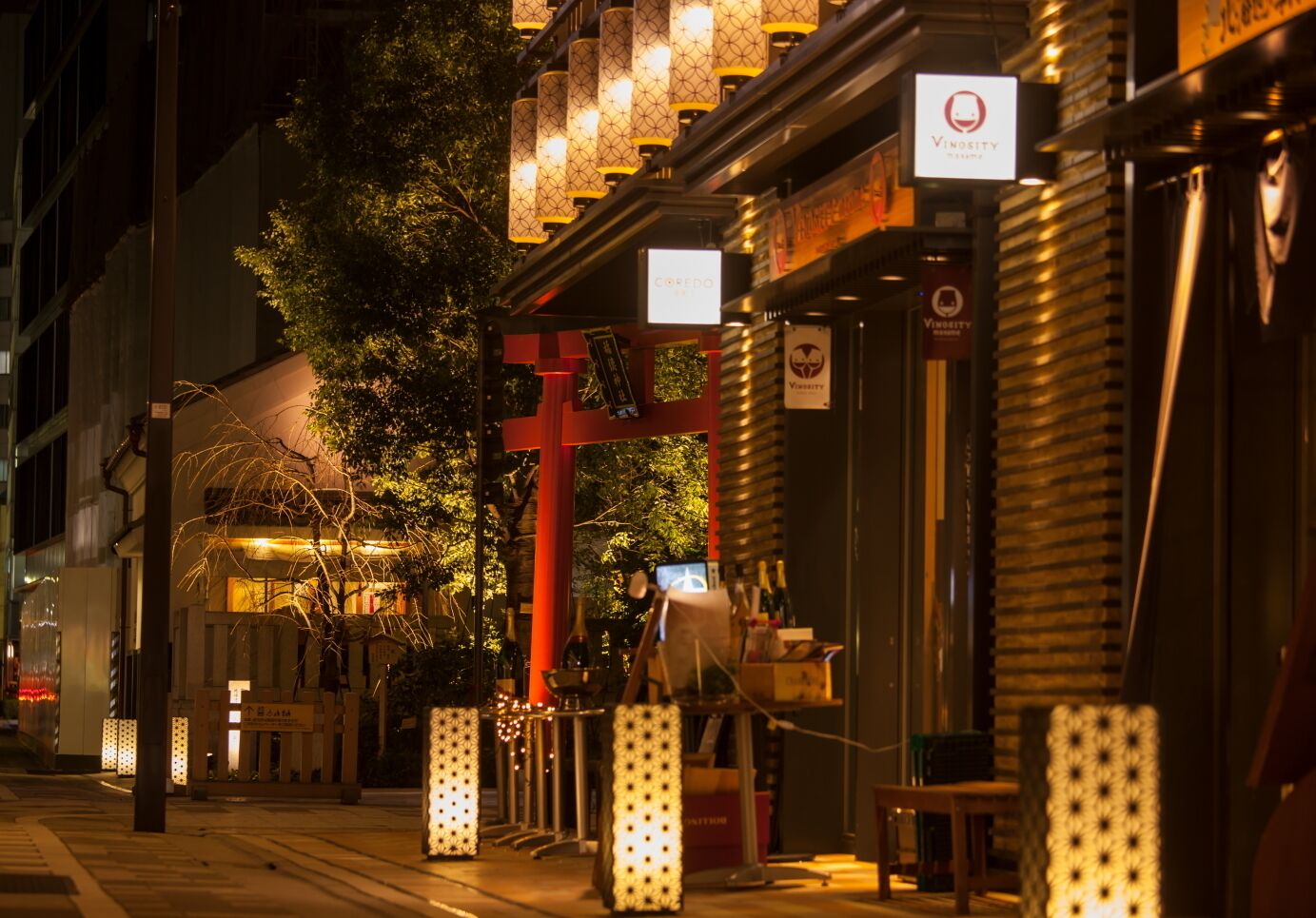 A street scene at night with a restaurant named 'Vinosity' and another named 'CREDO'. There are illuminated lanterns and a red torii gate.